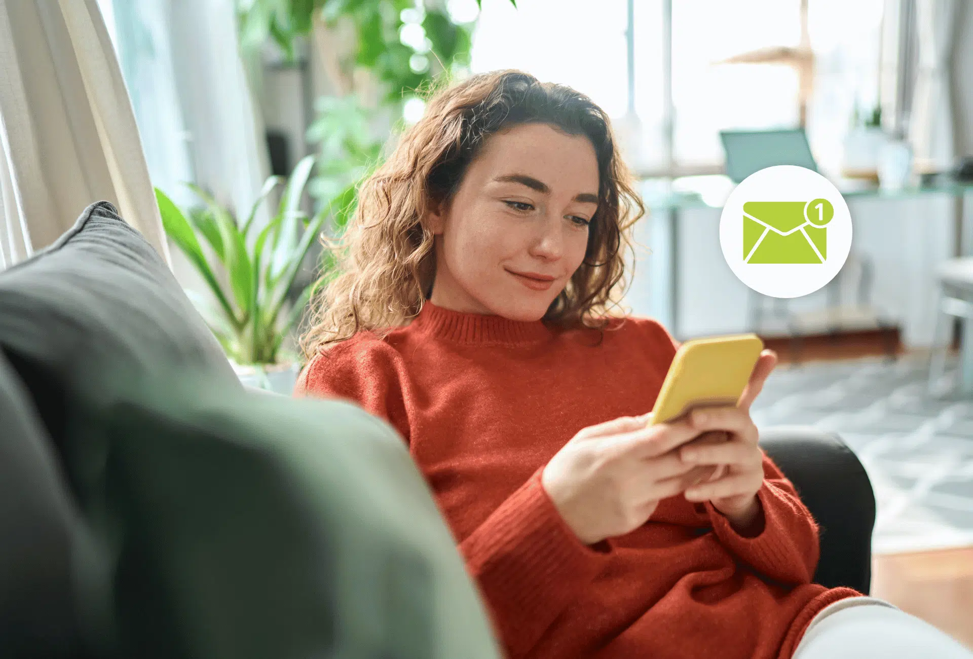 woman sitting on her couch in a bright room looking at her phone with an email icon overing above her phone